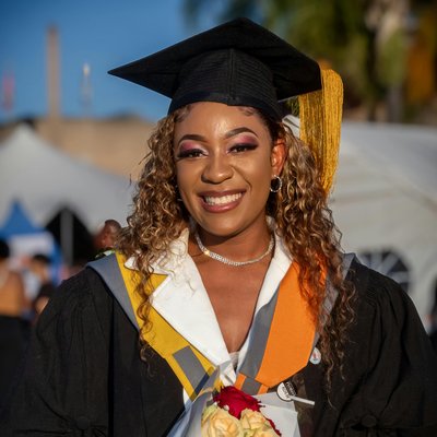 Woman in graduation regalia poses for a camera with a boquet of flowers in her hands.
